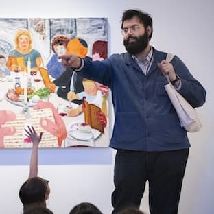 A museum educator stands in front of a colorful artwork depicting a lively dinner scene. The educator, wearing glasses, a beard, a blue jacket, and carrying a tote bag, gestures and points while engaging a group of children seated out of frame, one of whom raises their hand to ask a question.