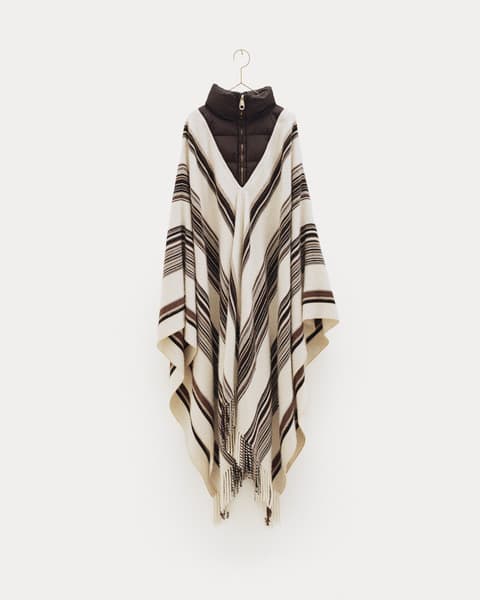 Beige poncho with brown and black horizontal stripe pattern and fringe hem, layered over a dark brown zip-up puffer vest with a high collar, displayed on a hanger against a white background.
