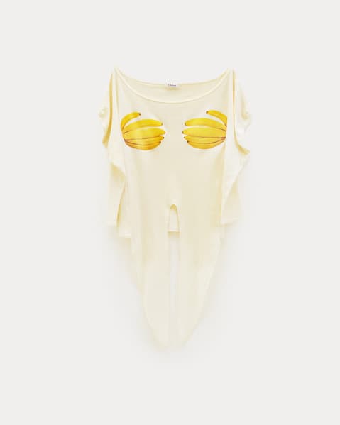 Off-white short-sleeved top with an asymmetrical, draped silhouette and high-cut center, featuring two stylized yellow banana graphics across the chest, displayed flat against a white background.
