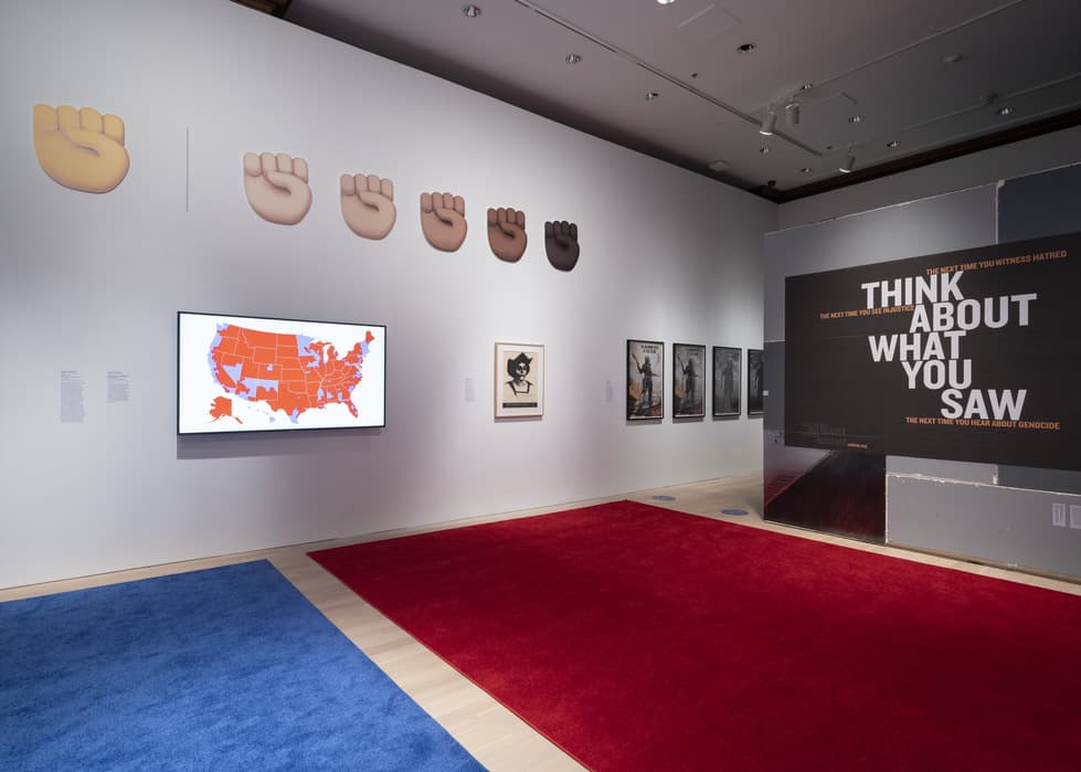 Gallery wall displaying a series of raised fist symbols in various skin tones, a U.S. map with red and blue states on a monitor, and framed artworks including a black-and-white portrait and several monochromatic prints. On the right, large wall text reads 