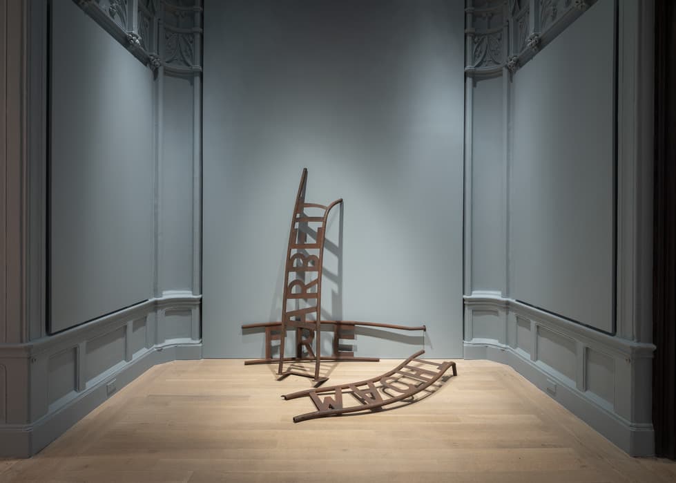 Metal sculpture of distorted gate fragments resting on the floor against a pale blue wall; one curved piece stands upright while others lie on the light wood floor, with the word 