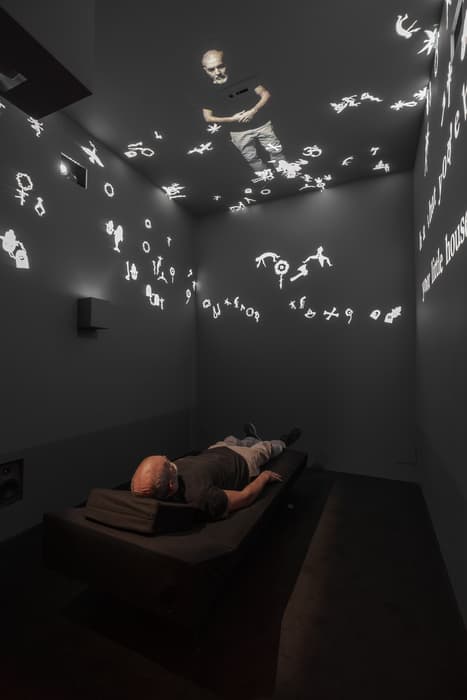 Man lying on a dark bench in a dimly lit room with symbols and a projection of himself illuminated on the ceiling and walls.