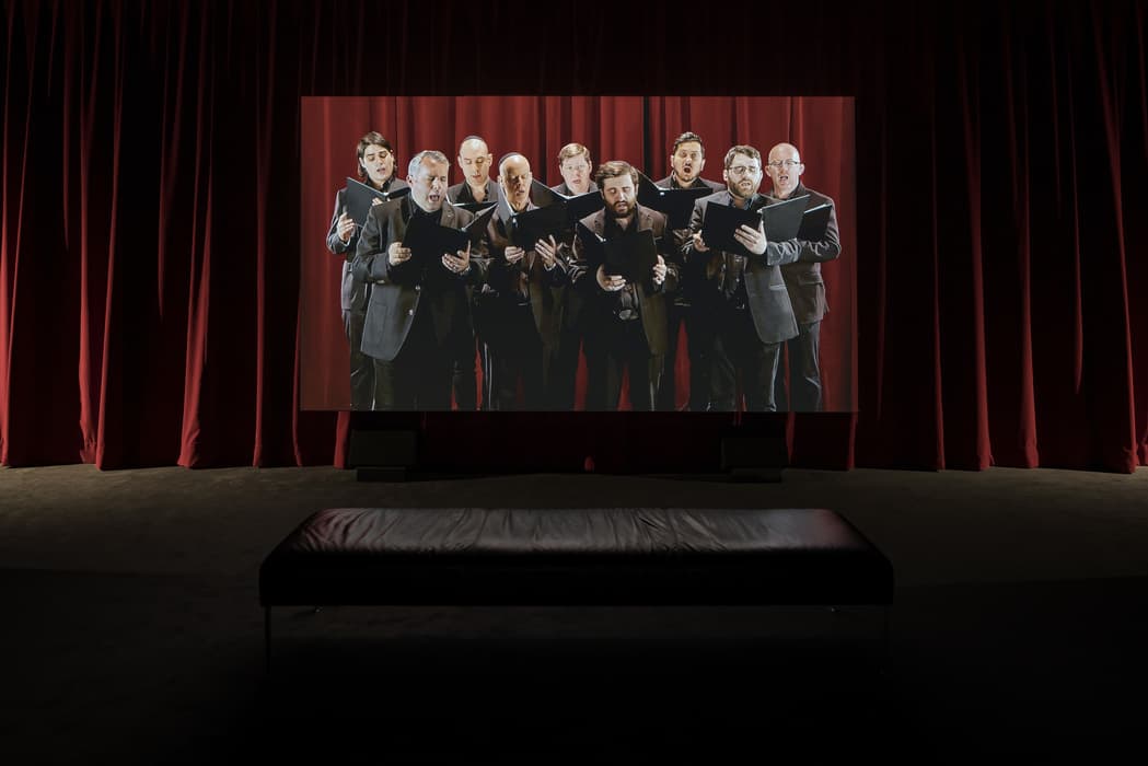 Video installation featuring a projection of a male vocal ensemble performing in front of red stage curtains, with a leather bench positioned for viewers in a darkened gallery space.