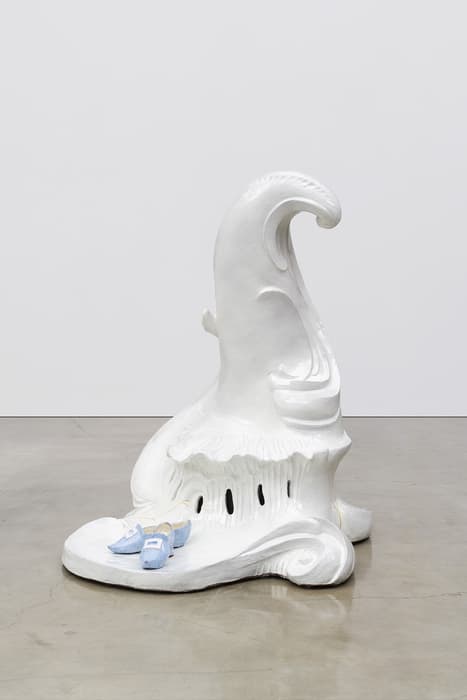 White sculptural form by Rachel Feinstein resembling a swirling, ornate rococo structure with a pair of small blue shoes placed at its base, set against a polished concrete floor and plain white wall.