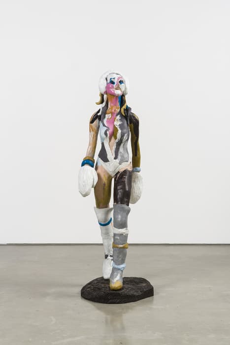 Sculpture of a female figure with exaggerated features, wearing a revealing bodysuit, large white earmuffs, mittens, and high boots, painted in a variety of bright colors and textures, standing on a dark base against a plain white background.