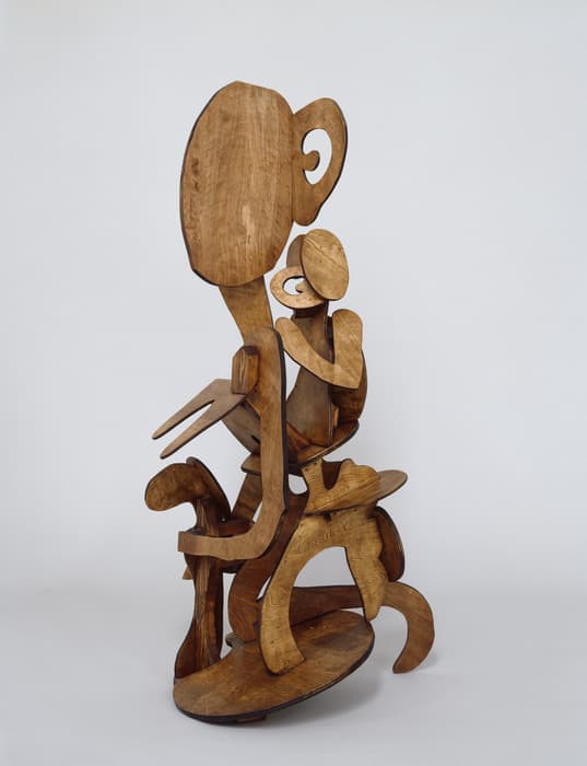 Abstract wooden sculpture composed of layered cut-out forms that suggest two stylized seated figures, one larger and one smaller, with oversized heads, simplified limbs, and curved organic shapes, all in a natural wood finish against a plain background.