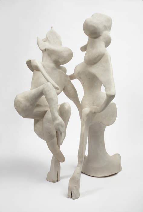 White sculptural piece featuring two abstracted, elongated seated figures with exaggerated, rounded facial features and limbs, posed in a dynamic, intertwined composition against a plain background.