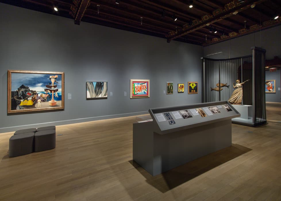 Exhibition gallery with paintings and sculptures on view; artworks include colorful abstract and figurative pieces on dark gray walls, with a central display case holding archival materials.