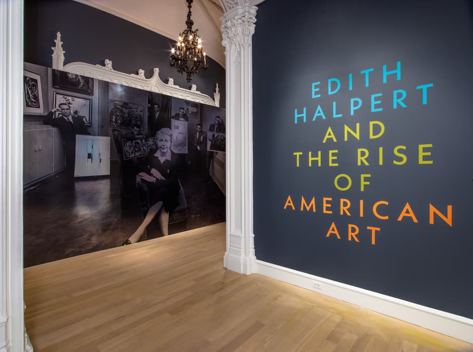 Exhibition entrance with large text reading “Edith Halpert and the Rise of American Art” on a dark blue wall; adjacent is a black-and-white photo mural of Edith Halpert seated with artists and artworks.