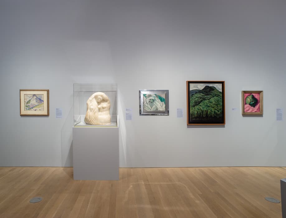 Gallery wall displaying five works: four framed paintings of landscapes and flowers, and a central white stone sculpture of a seated figure under a glass case, all on light gray walls with wood flooring.