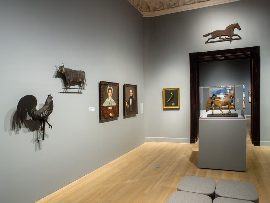Gallery with folk art sculptures and paintings: metal rooster and cow weathervanes on the left wall, two framed portraits and a small framed firearm, a wooden horse sculpture in a glass case, and a running horse silhouette above a doorway.