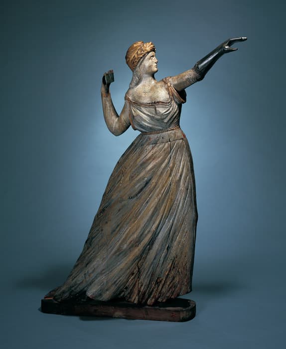 Carved wooden ship figurehead of a woman in flowing robes, one arm extended forward and the other raised, wearing a crown or headdress.
