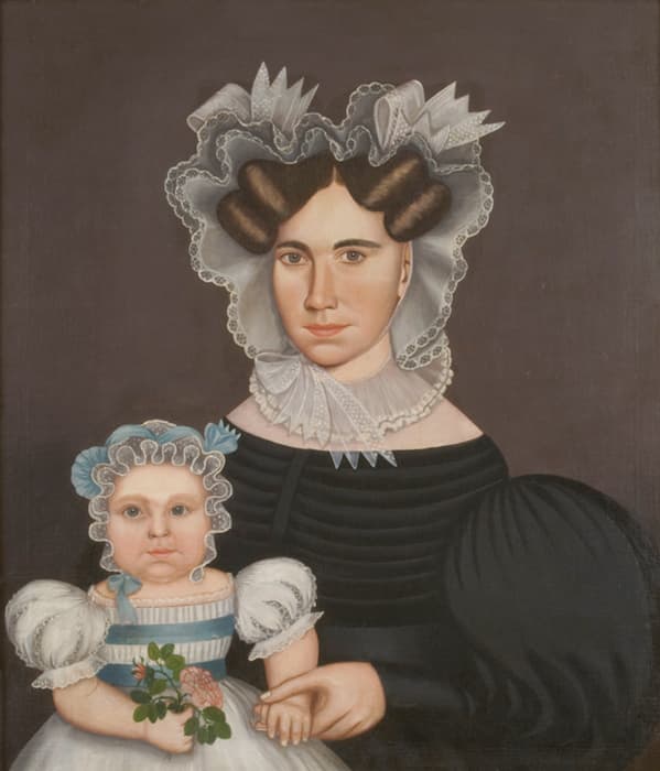 Portrait of a woman in a black dress with elaborate lace bonnet holding the hand of a child in a white dress and bonnet, who holds pink flowers.