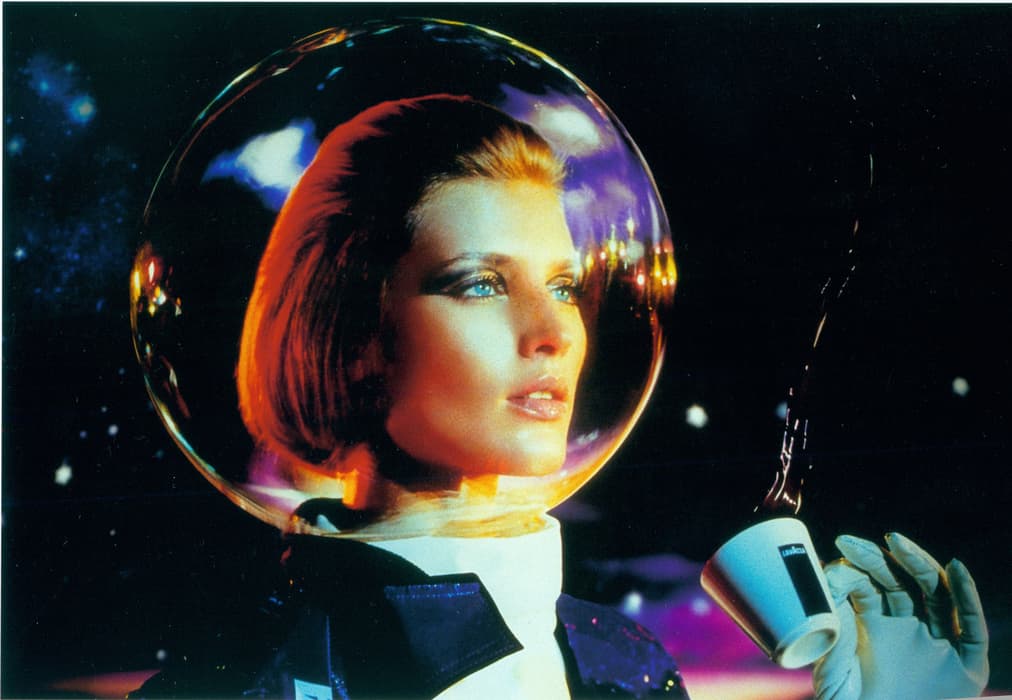 A woman in a futuristic spacesuit with a transparent bubble helmet gazes ahead while holding a small espresso cup mid-pour in a surreal, outer space-like setting filled with colorful reflections and stars.