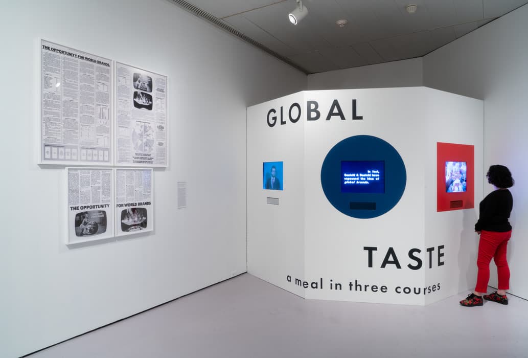 Art exhibition corner labeled “GLOBAL TASTE: a meal in three courses” features three built-in screens displaying video content. A visitor in red pants watches one of the screens. On the adjacent wall, framed newspaper-style posters titled “THE OPPORTUNITY FOR WORLD BRANDS” are displayed, discussing advertising and globalization.