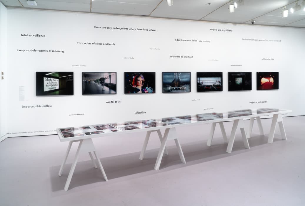 A contemporary art gallery installation featuring a white wall with mounted digital screens displaying various images, including a woman in a space helmet and airport scenes. Phrases and poetic text fragments such as 