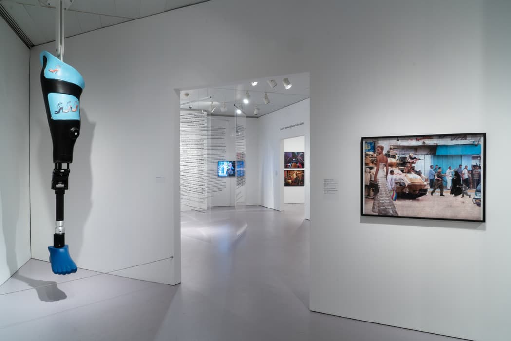 Contemporary art gallery with a large, blue prosthetic leg hanging from the ceiling on the left and a framed photo collage on the right wall showing a woman in a dress walking past a military scene. Through the doorway, suspended transparent panels with printed text and additional video and photographic artworks are visible in an adjacent gallery room.