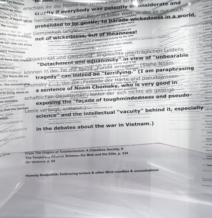 Close-up view of a transparent installation panel layered with overlapping English and German text in various sizes and fonts. Phrases like “Detachment and ‘equanimity’ in view of ‘unbearable tragedy’ can indeed be ‘terrifying’” and “in the debates about the war in Vietnam” are visible. Citations at the bottom reference works by Hannah Arendt and others. The background shows a faint gallery space with white walls and minimal lighting.
