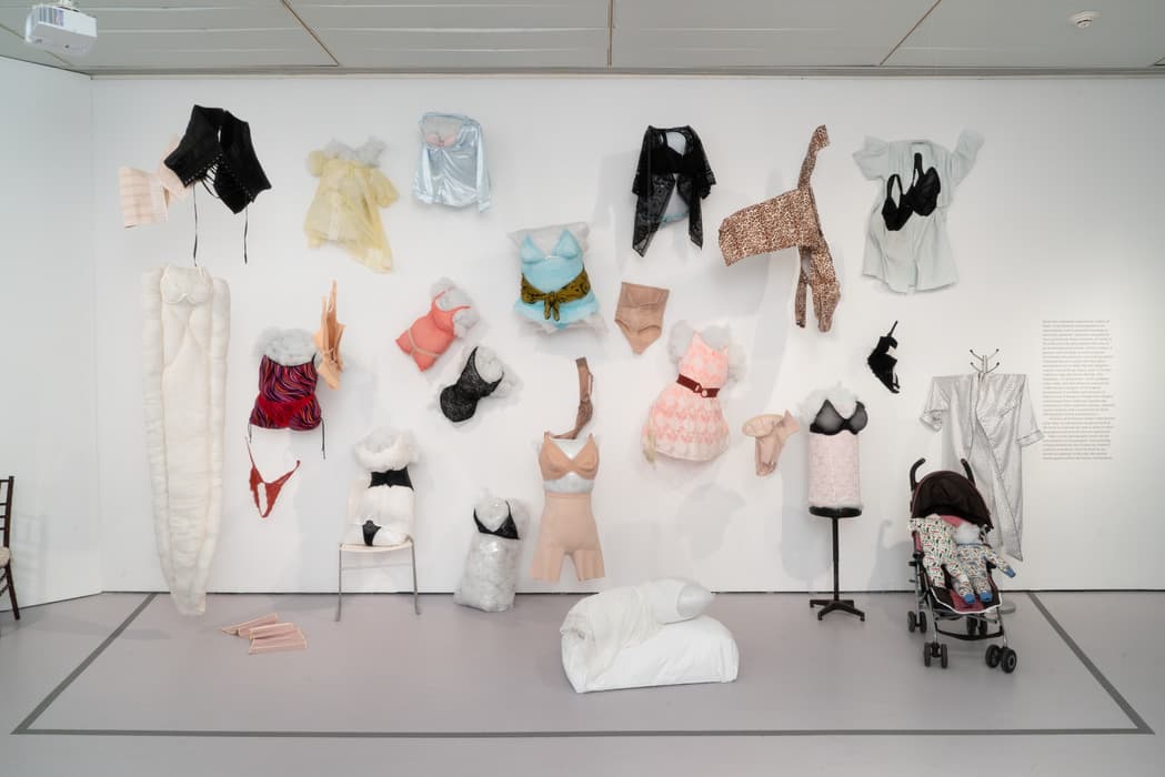 Art installation of women’s clothing and intimate wear displayed on a gallery wall, including bras, nightgowns, and shapewear arranged on pillows, mannequins, and hangers. A white padded sleeping bag, a child-sized mannequin in a stroller, and a soft sculpture on the floor accompany the wall display. A text panel is visible on the right side.