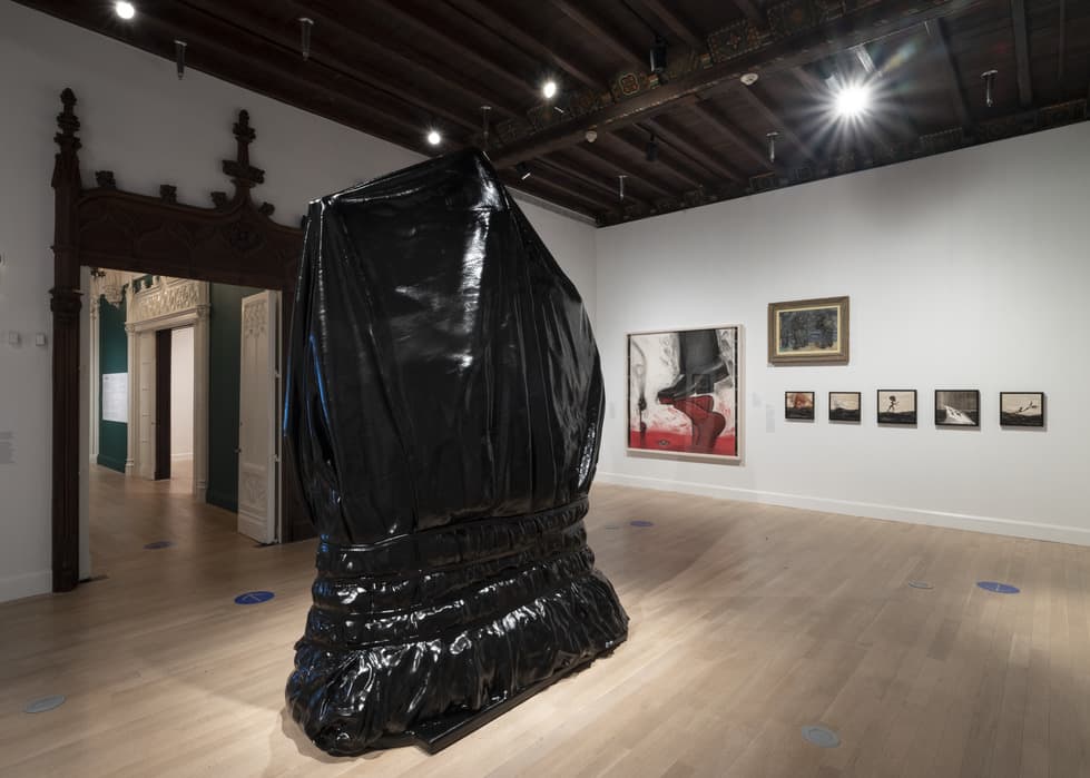 Gallery interior featuring a tall, glossy black draped sculpture centered on light wood flooring; background wall displays five framed artworks, including a large abstract piece with red and black tones and four smaller photographs; ornate dark wood entryway visible to the left, leading into an adjacent room.