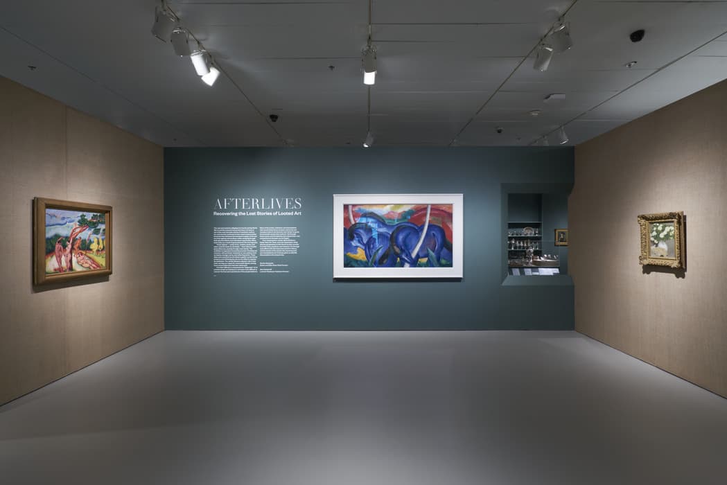 Gallery installation view of Afterlives: Recovering the Lost Stories of Looted Art. Three framed paintings hang on the walls, including a colorful depiction of blue animals at center, flanked by works in ornate gold frames on textured tan walls. Exhibition text is displayed on the central dark teal wall. A recessed display of silver objects is partially visible to the right.