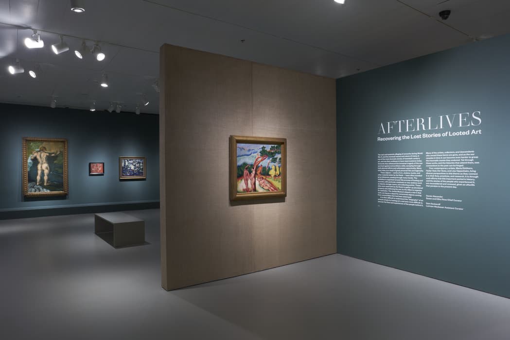 Gallery installation view of Afterlives: Recovering the Lost Stories of Looted Art. Exhibition text is displayed on a dark teal wall at right, next to a freestanding tan partition holding a framed painting of reclining nudes beneath a tree. In the distance, additional artworks hang on dark teal walls, including a large full-length nude figure at left and two smaller paintings. A bench sits in the middle of the gallery space under bright ceiling track lights.