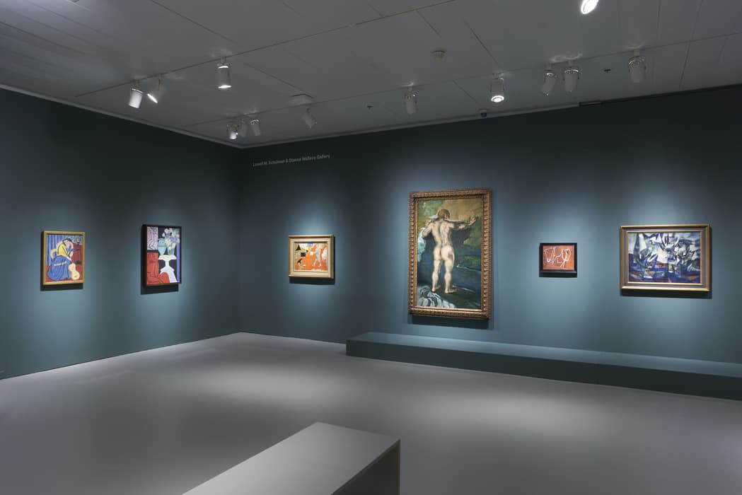 Gallery installation view showing six framed paintings hung on dark teal walls under spotlights. From left to right: a seated figure with a guitar in vibrant colors, a still life with flowers, a small orange and white figurative scene, a large full-length painting of a nude figure seen from behind, a small abstract line drawing of figures, and a Cubist-style composition with angular shapes. A low bench and platform sit in the center of the minimalist gallery space.