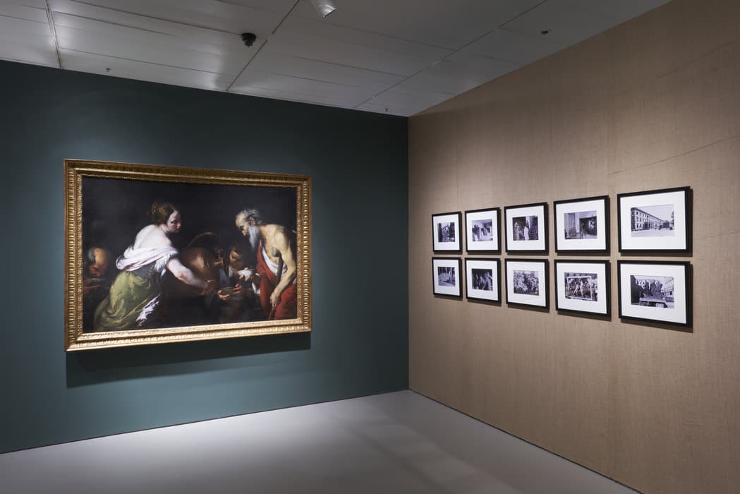 Gallery installation view featuring a large, framed oil painting on the left wall, depicting a biblical or historical scene with multiple figures, including a woman pouring water and an elderly man drinking. The painting is displayed on a dark green wall. To the right, on a burlap-textured wall, are two rows of small framed black-and-white photographs documenting various scenes of people, architecture, and events. The space is minimal, with neutral flooring and ceiling spotlights.