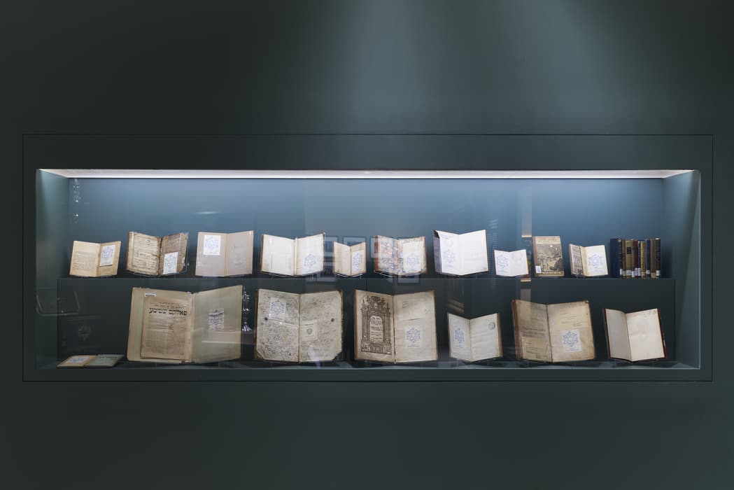 Glass display case with two shelves of open books and manuscripts, some showing Hebrew text, illuminated in a dark teal wall. A variety of historical book covers, pages, and bindings are visible, including a few books standing closed on the right.