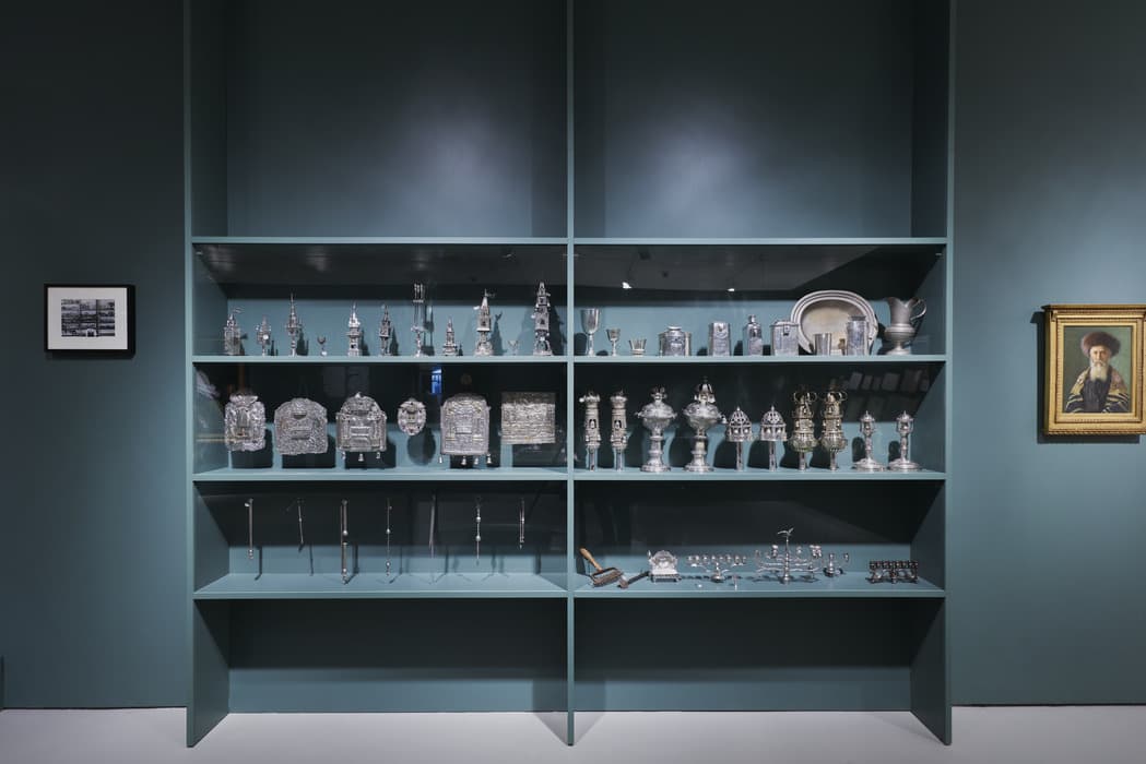 Display case with multiple shelves showcasing a collection of silver Judaica objects, including Torah crowns, finials, breastplates, kiddush cups, spice towers, menorahs, and other ceremonial items. The shelves are built into a dark teal wall. To the left of the display is a small black-and-white photograph, and to the right hangs a framed portrait of a man with a beard and traditional fur hat, dressed in ceremonial robes.