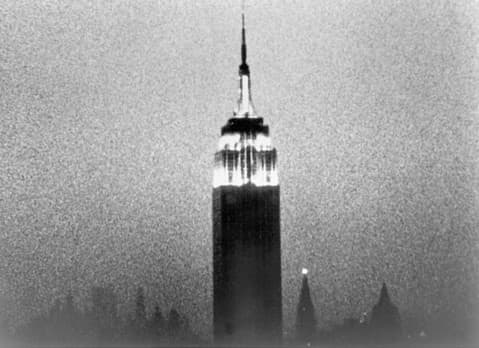 Grainy black and white photograph of the Empire State Building at night, with its top brightly illuminated against a hazy, dark sky. The silhouette of other New York City buildings is faintly visible at the bottom of the image.
