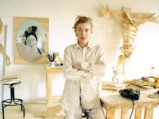 Artist Rachel Feinstein stands in her studio, wearing a white textured outfit covered in paint. Behind her are wooden sculptures, tools, and a workbench with materials.