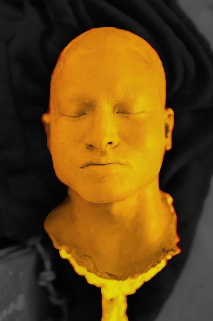 Sculptural bust of a human head with closed eyes, painted in bright yellow-orange, set against a dark black fabric background.