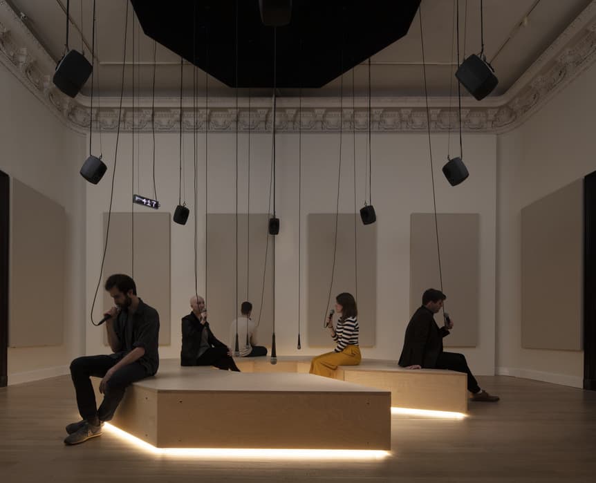 Immersive sound installation with visitors seated on illuminated wooden benches, each holding a microphone, surrounded by suspended speakers in a dimly lit gallery space.