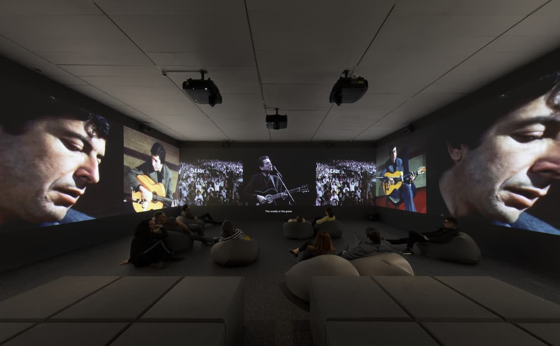 Immersive video installation with multiple large screens displaying close-ups and performances of Leonard Cohen, surrounded by seated viewers on bean bags in a darkened room.