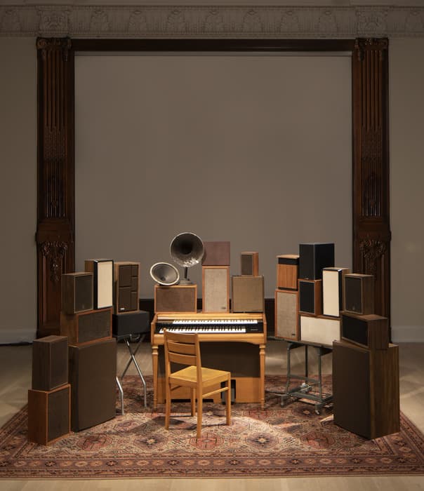 Wooden chair and organ surrounded by vintage speakers and gramophones arranged on an ornate rug within a classical architectural frame.