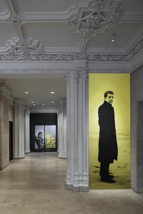 Museum entrance with ornate white columns and two large photographic posters of Leonard Cohen, one in black and white and one in yellow tones.
