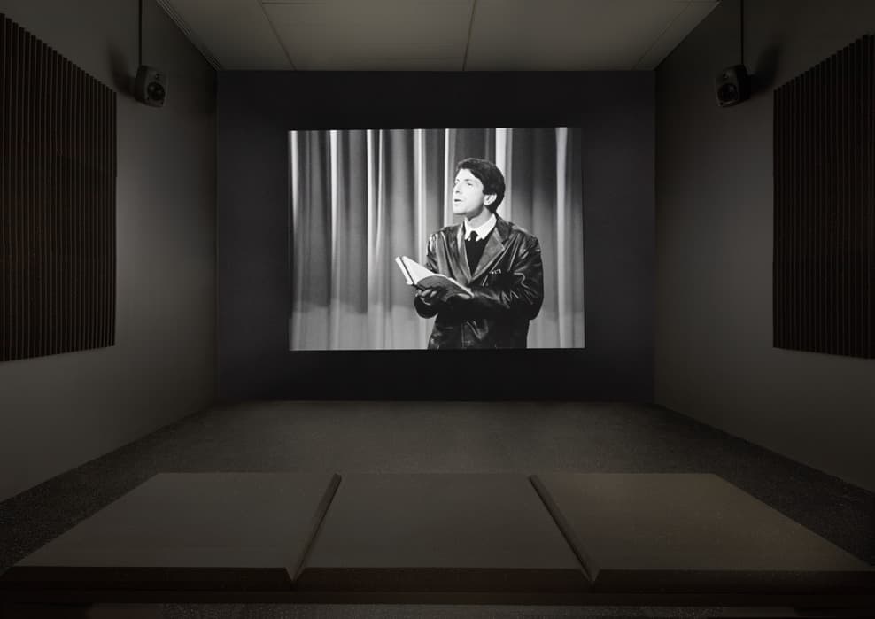 Black-and-white video installation in a dark gallery showing a young man reading from a book, projected on a screen with seating cushions in the foreground.
