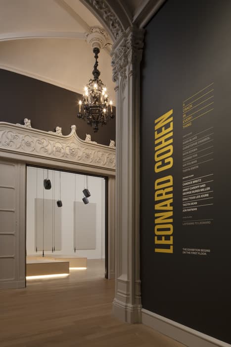 Exhibition entrance with ornate architectural details, a chandelier, and a wall featuring the name “Leonard Cohen” in bold yellow text alongside a list of exhibition sections.
