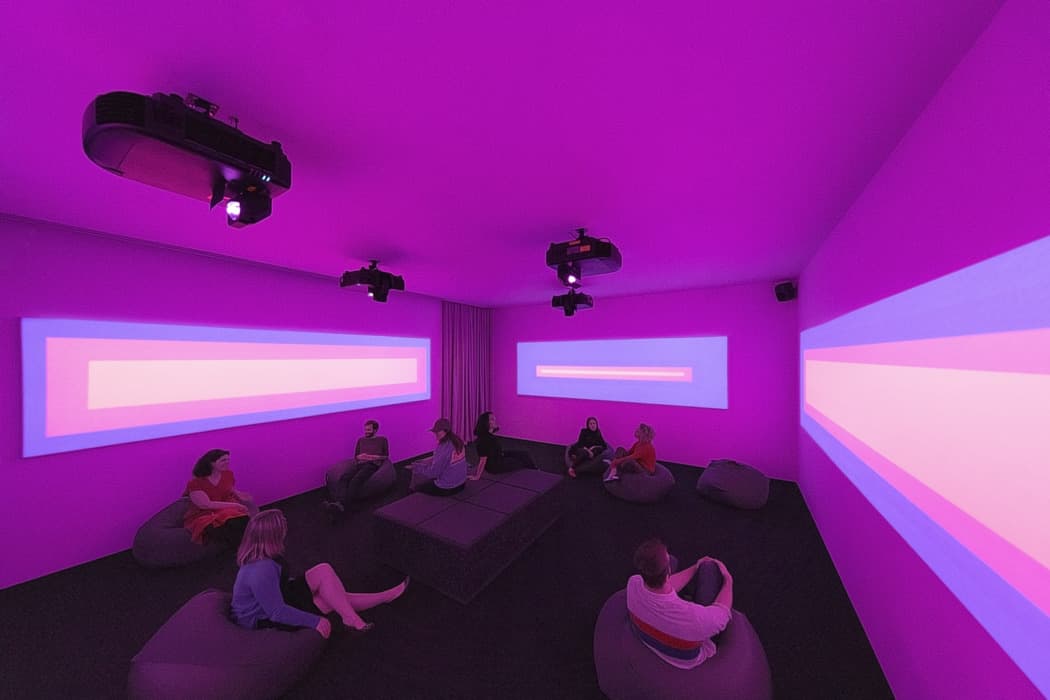 Immersive purple-lit room with abstract geometric projections on three walls, where visitors sit on bean bags watching the installation.