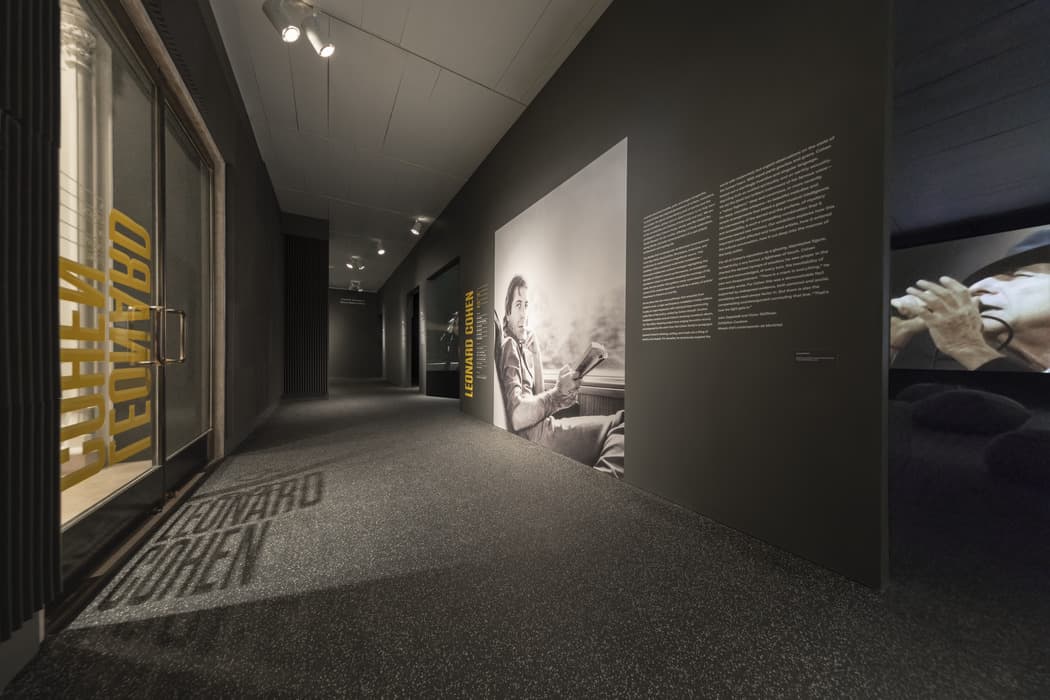 Hallway of a Leonard Cohen exhibition featuring a large black-and-white photo of Cohen reading on a train, adjacent to explanatory wall text and projected visuals.