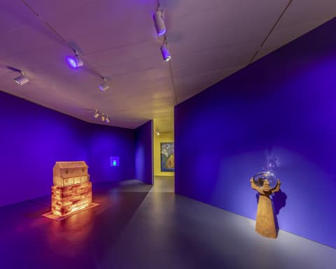 Exhibition gallery with deep blue walls and dramatic lighting, featuring a glowing wooden sculpture resembling a chest on a brick base to the left and a brown sculpture holding a reflective orb to the right, with additional artwork visible in a yellow-lit room beyond.