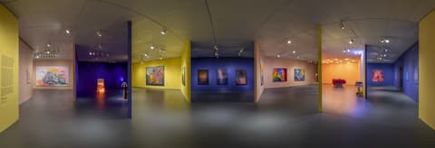 Panoramic view of a contemporary art gallery with multiple rooms painted in yellow, blue, and peach tones, displaying colorful abstract paintings, sculptures, and installations under track lighting.