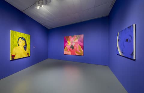 Gallery room with deep blue walls displaying three contemporary portraits: a yellow-toned painting of a young girl on the left, a pink abstract figure in the center, and a close-up blue portrait of a face on the right.