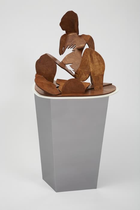 Wooden cut-out sculpture of a stylized seated female figure, composed of flat shapes with curved edges, displayed on a tall gray pedestal with a white rim.