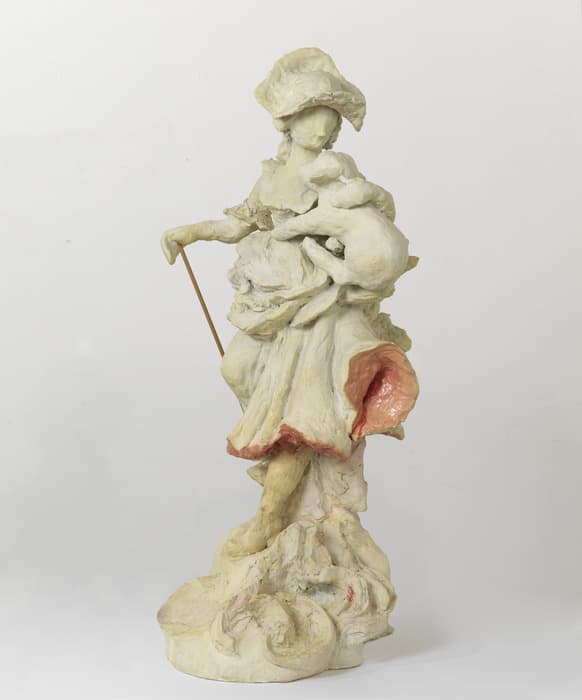 Sculpture of a shepherdess figure with a bonnet and staff, cradling a lamb, rendered in pale, textured material with pink accents inside the skirt and at the base.