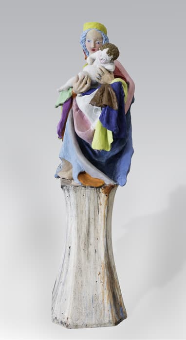 Colorful sculpture of a seated woman holding a child, both wearing draped, multicolored garments, positioned on top of a tall, weathered pedestal.