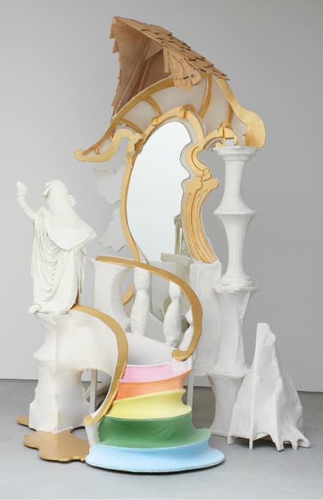 Large whimsical sculpture resembling an ornate vanity with a central mirror, curved gold and white architectural elements, rainbow-colored steps, and a small white robed figure gesturing upward.