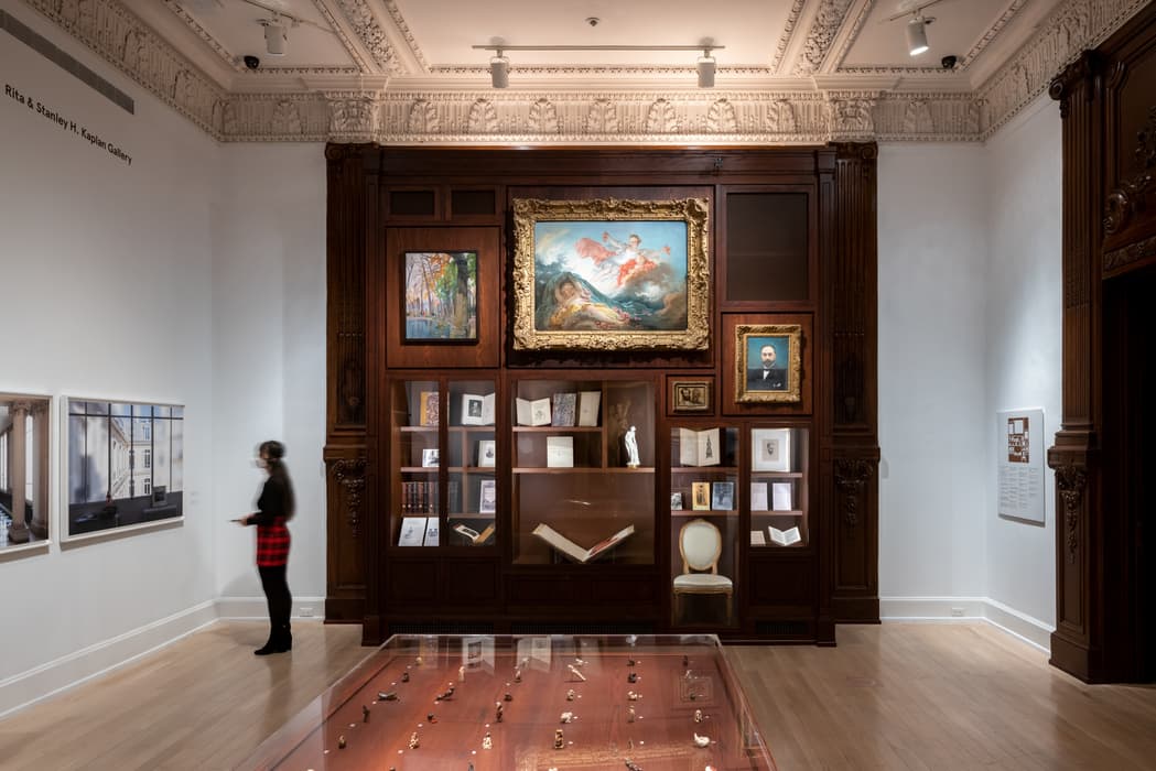 Gallery view with display case of small objects in foreground, books and artwork arranged in a large wooden cabinet with framed paintings above.