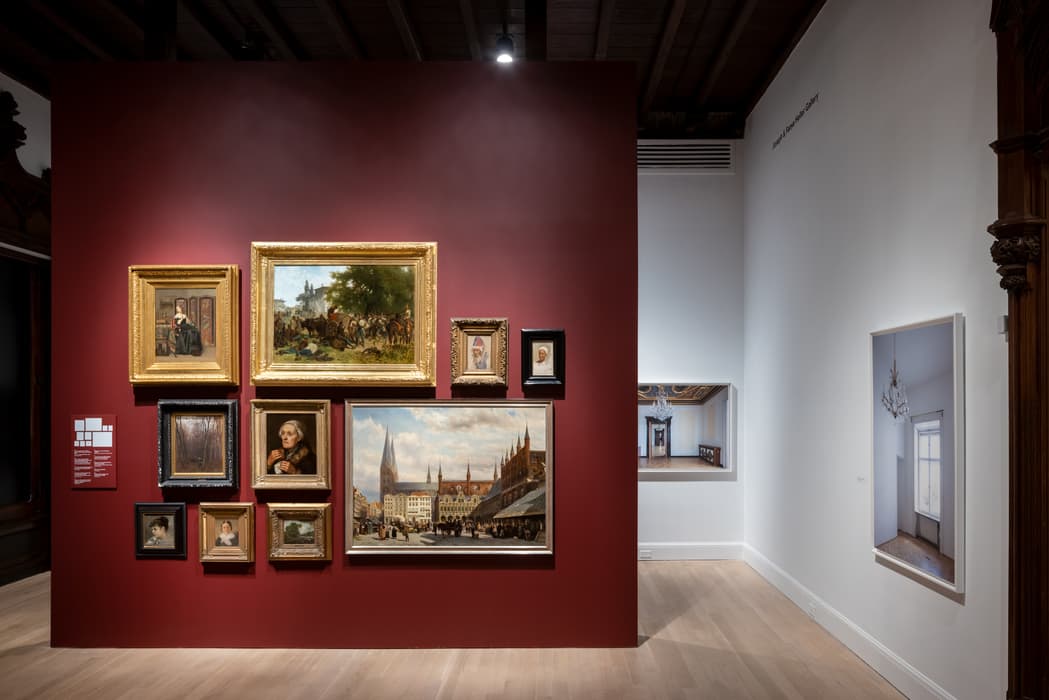 Gallery view with a red accent wall displaying a salon-style arrangement of framed paintings, including portraits and cityscapes; additional photographs hang on the adjacent white wall.